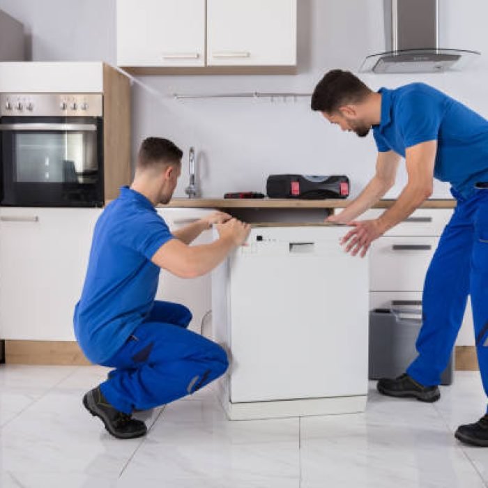 Two Young Male Movers In Uniform Placing Dishwasher In Kitchen Two Young Male Movers In Uniform Placing Dishwasher In Kitchen